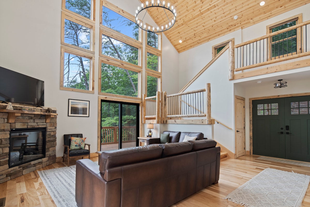 A spacious living area features tall windows allowing natural light to flood in, highlighting the wooden beams and cozy seating arrangements. A stone fireplace adds a rustic touch, while the open staircase leads to the upper level, enhancing the inviting feel of the cabin.