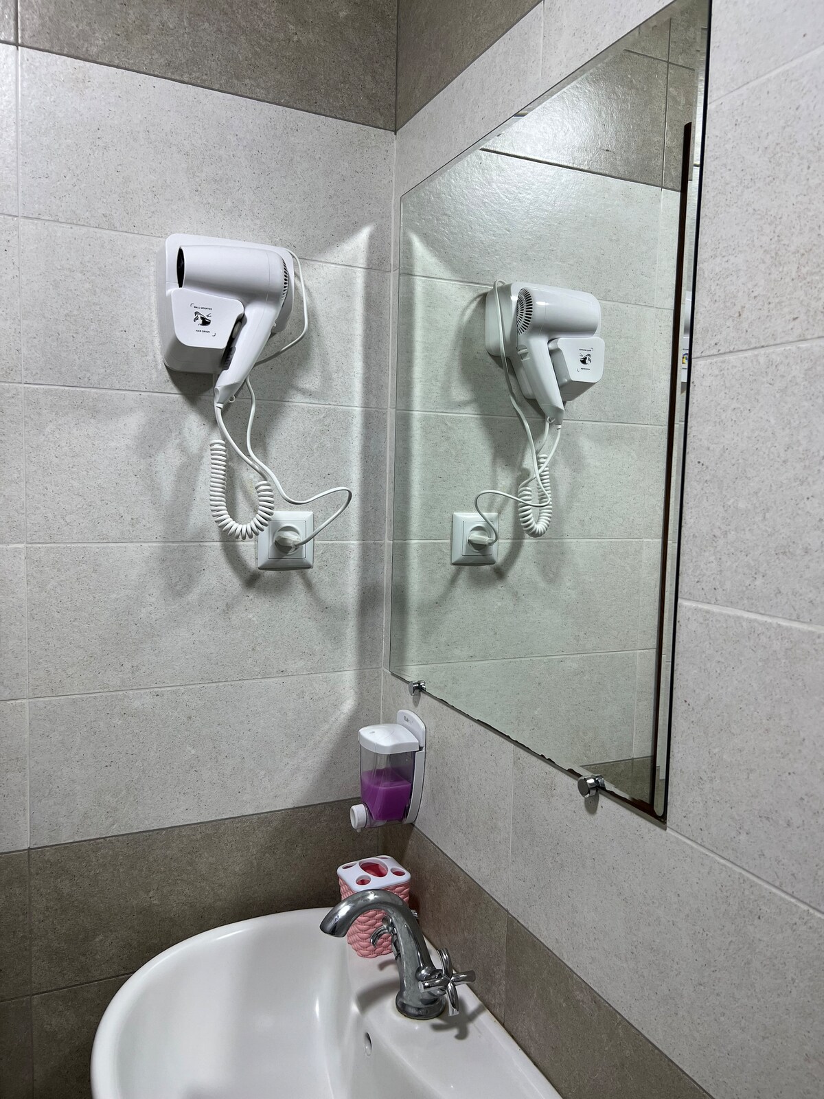 A modern bathroom features smooth tile walls in neutral tones. Two hair dryers are mounted beside a large mirror, reflecting the well-lit space. A small dispenser of liquid soap is attached to the wall, positioned above a white sink with a sleek faucet.