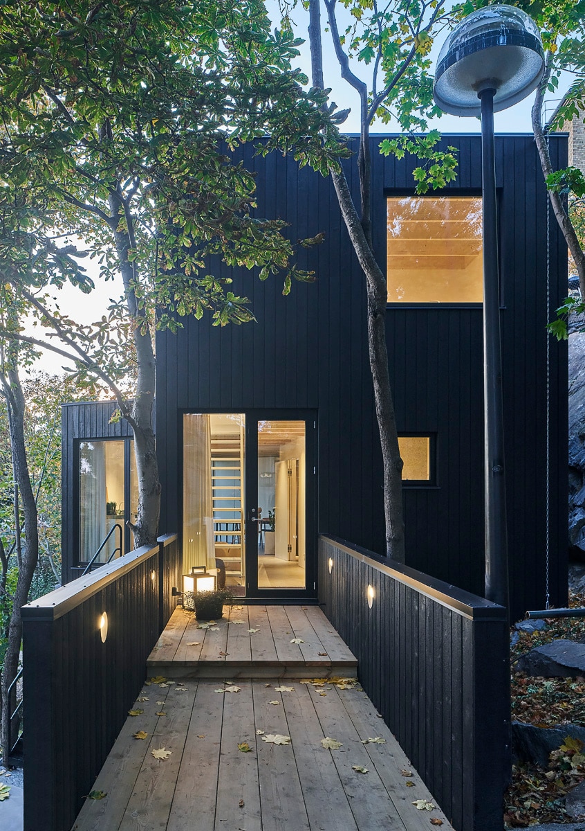 The entrance to The Charred House is depicted, showcasing a modern black exterior surrounded by trees. A wooden walkway leads up to the front door, illuminated by soft lighting. Large windows provide glimpses of the interior, enhancing the connection with nature.