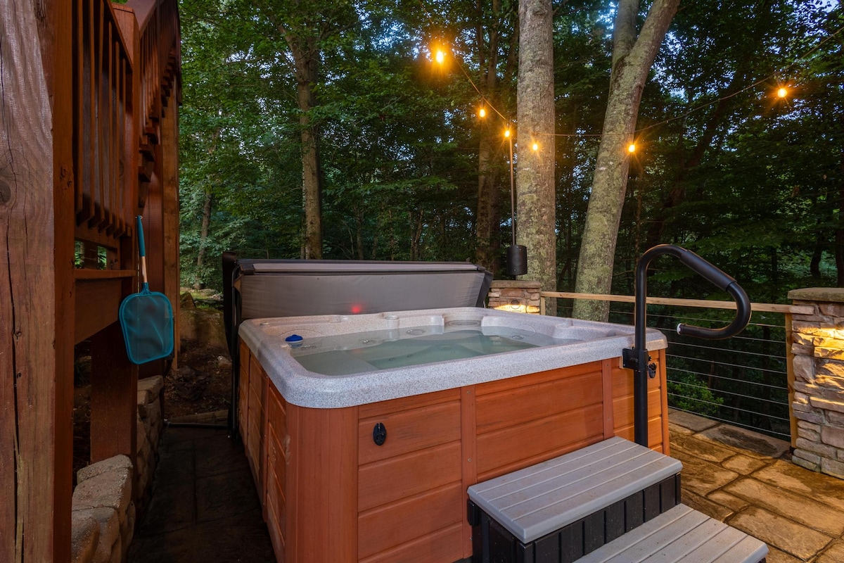 A private hot tub is positioned on a stone patio, surrounded by trees and softly illuminated by string lights. The area features a sturdy railing, enhancing the secluded outdoor setting.