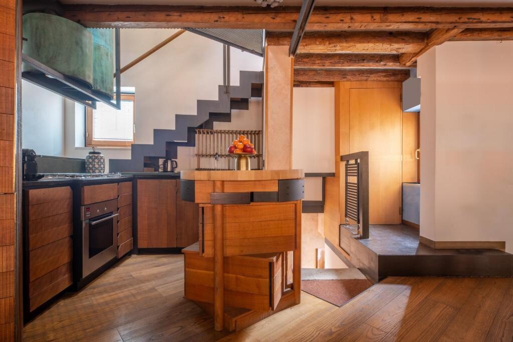 The open kitchen features modern appliances and wooden cabinetry, complemented by a round dining table with matching chairs. A staircase leads to the upper levels, with wooden beams visible on the ceiling. Natural light enters from a nearby window.