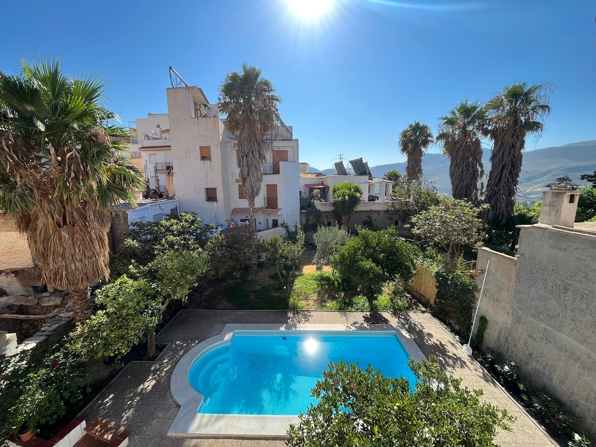An aerial view captures a private pool surrounded by lush greenery and palm trees. The sun shines brightly overhead, illuminating the garden and the charming layout of the property. The residential structure and surrounding area are visible in the background.
