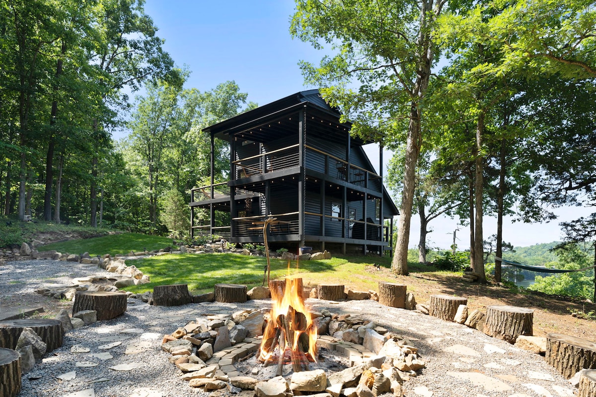 A modern cabin is surrounded by trees, featuring multiple levels and outdoor decks. A fire pit made of stone and wood logs is centered in a gravel area, inviting guests to enjoy the peaceful woodland setting.