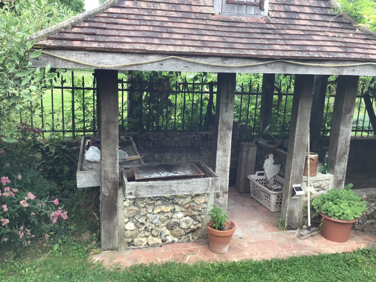 An outdoor barbecue area is situated under a wooden shelter with a sloped roof. Stone and wood elements create a rustic feel. Potted plants and flowers are visible around the patio, enhancing the natural surroundings. The area appears well-maintained and inviting for outdoor cooking.