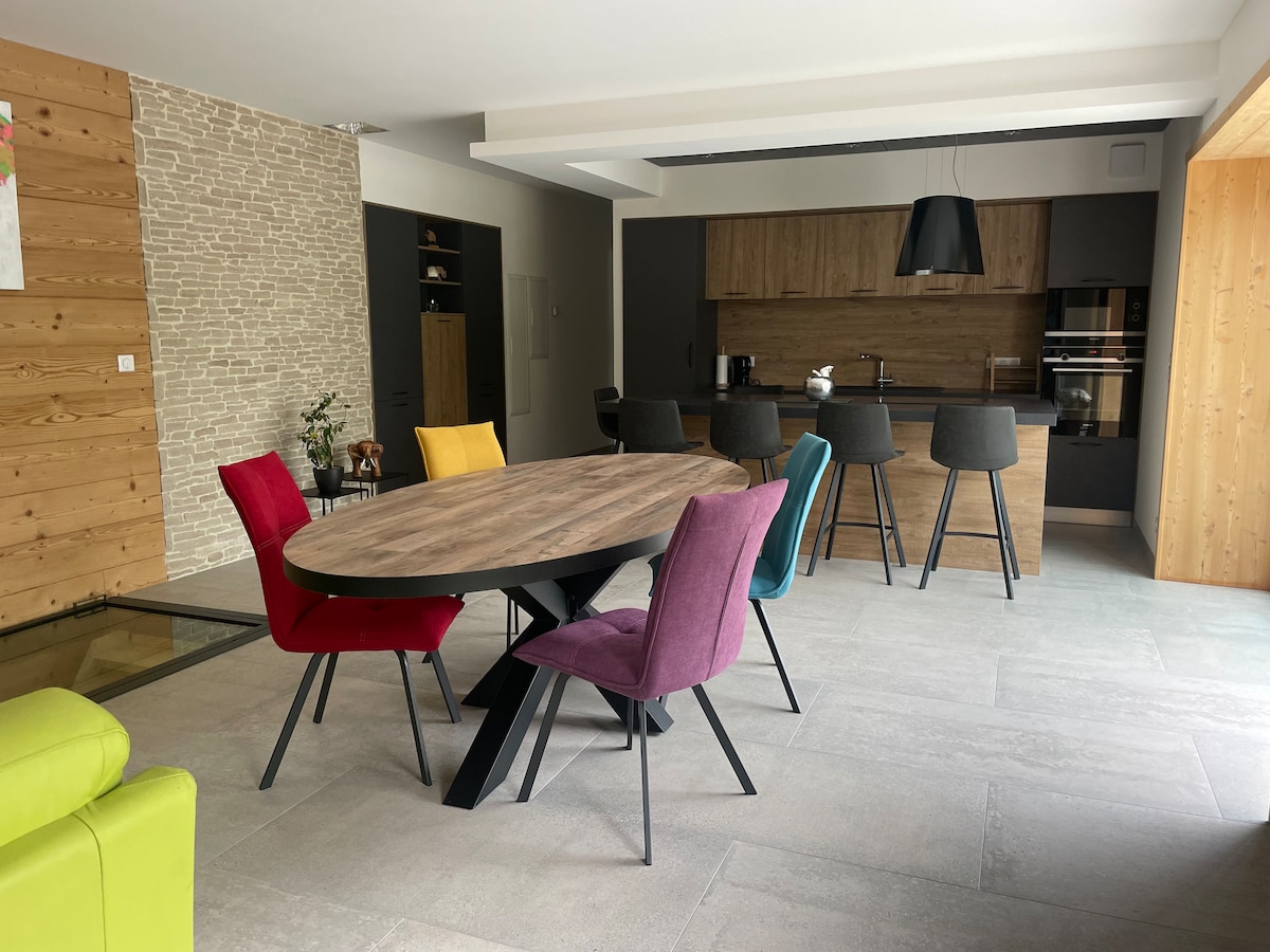 A modern dining area is presented, featuring a round wooden table surrounded by colorful upholstered chairs in red, purple, and yellow. A sleek kitchen with dark cabinetry is visible in the background, complemented by stone and wooden accents on the walls.