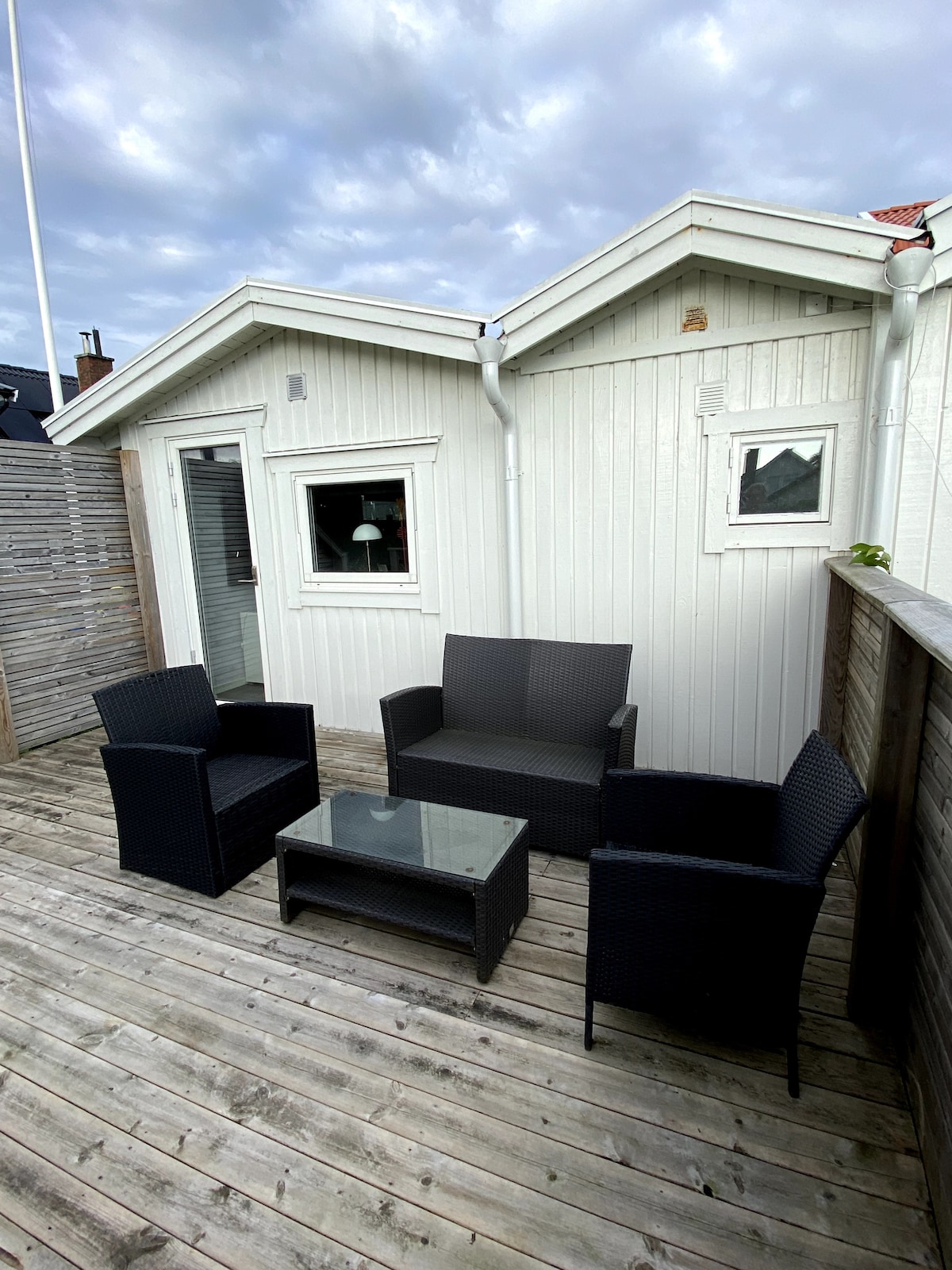 An outdoor balcony area features a seating arrangement with four dark wicker chairs and a glass-topped coffee table. The wooden deck showcases natural tones, while the white exterior walls of the nearby structure provide a contrasting backdrop.
