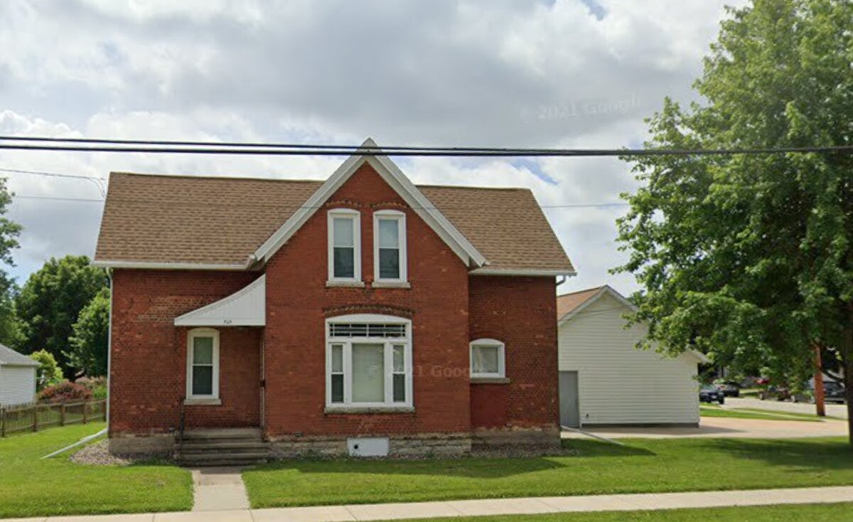 The exterior of a two-story red brick house is visible, featuring a gabled roof and a front porch with steps leading up. The façade includes white-framed windows, and a nearby grassy area is bordered by trees. A garage is seen to the side.