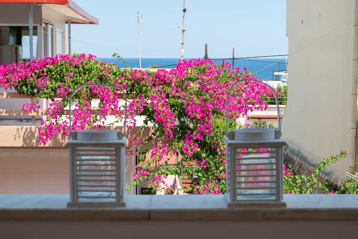 A balcony view showcases vibrant bougainvillea in full bloom with rich pink flowers. Two lanterns sit on the railing, framed by the colorful plant life. The ocean is visible in the distance, blending seamlessly with the clear sky.