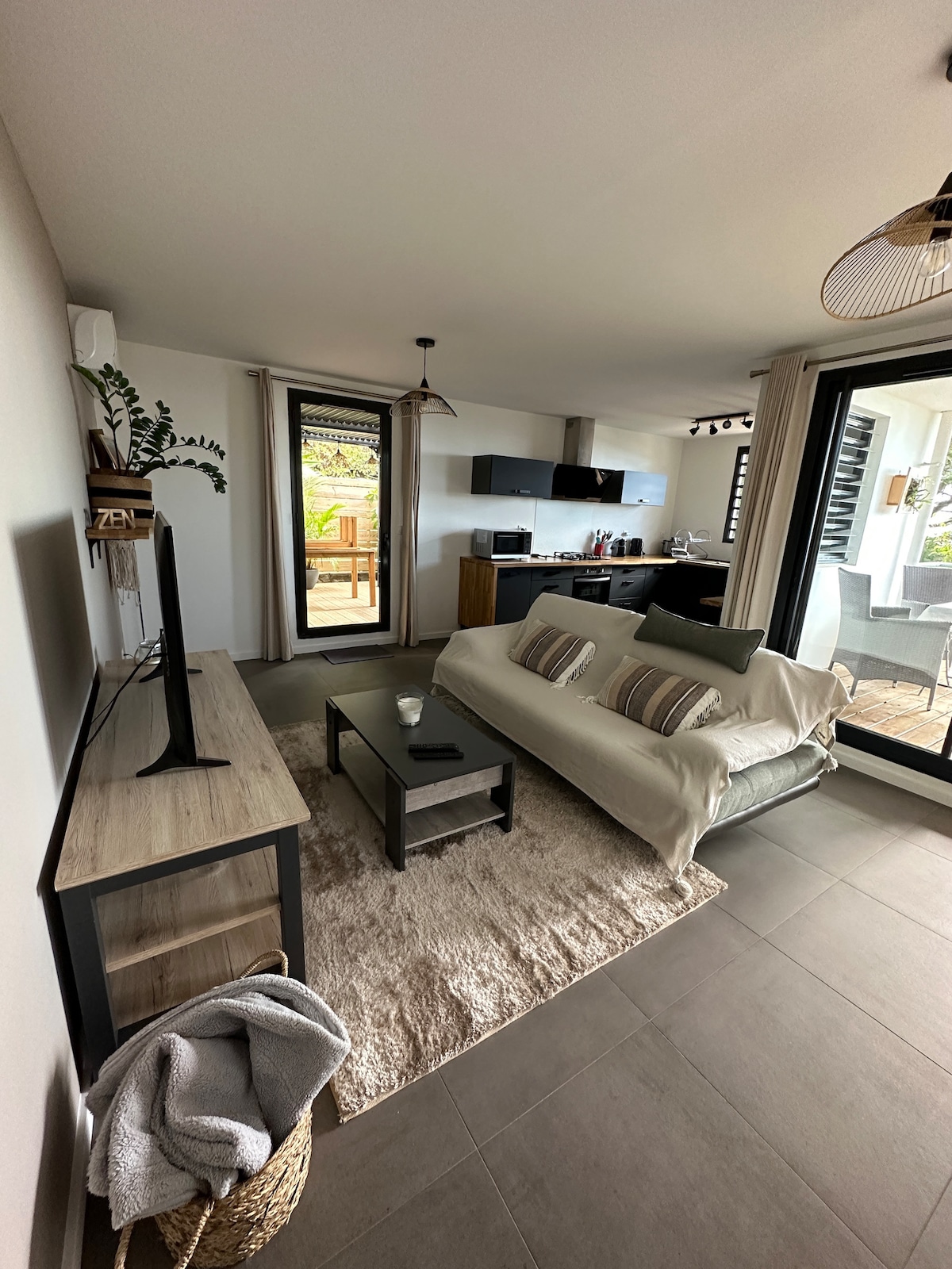 A modern living space features a light sofa with decorative pillows, a coffee table, and a flat-screen TV on a wooden stand. Large windows provide access to an outdoor area, while a rug adds comfort to the tiled floor.