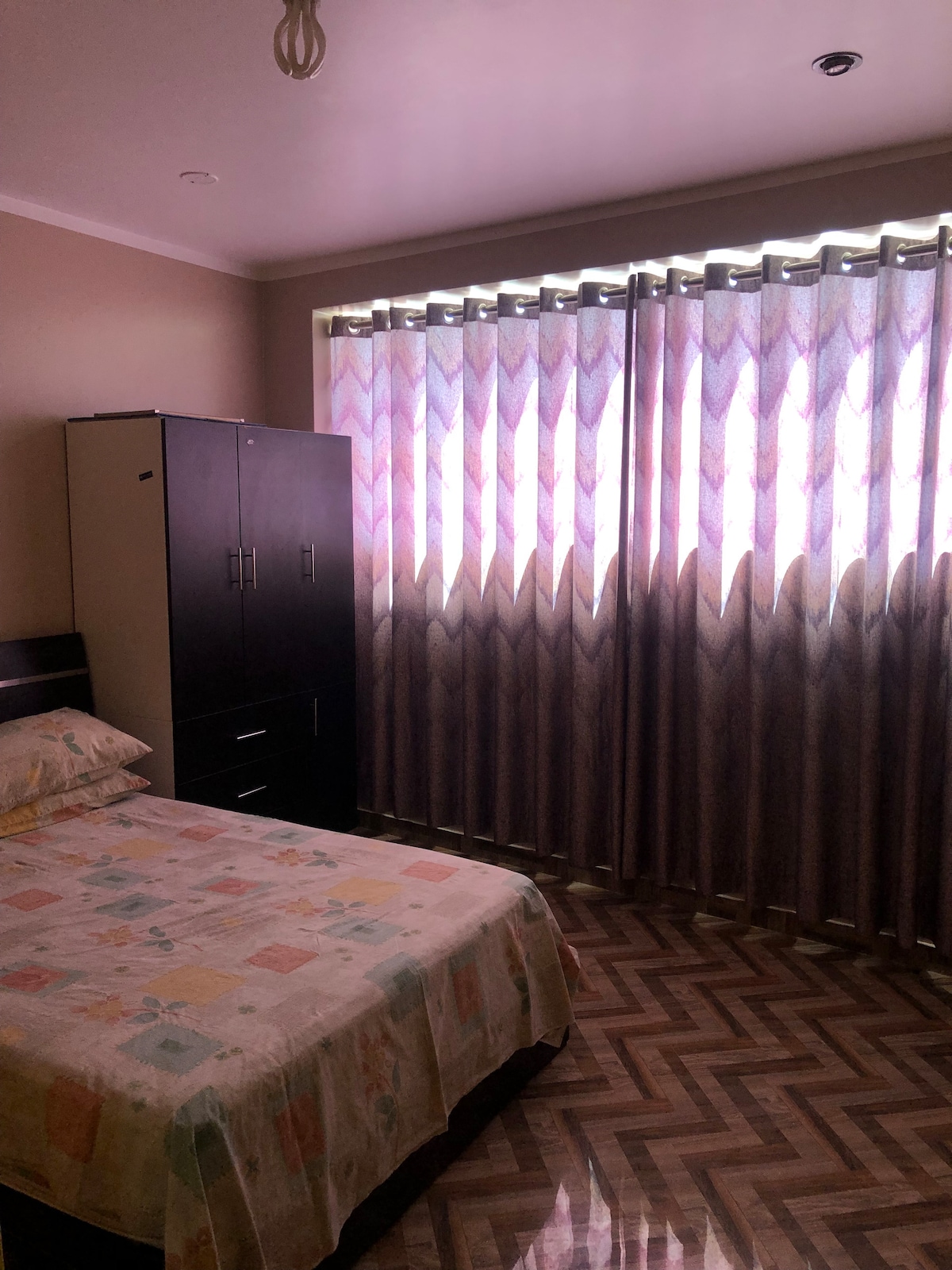 A cozy room is depicted featuring a spacious bed covered with a patterned bedspread. A large wardrobe stands next to the bed, while pull-down blinds allow natural light to filter through, creating a soft ambiance in the well-lit space.