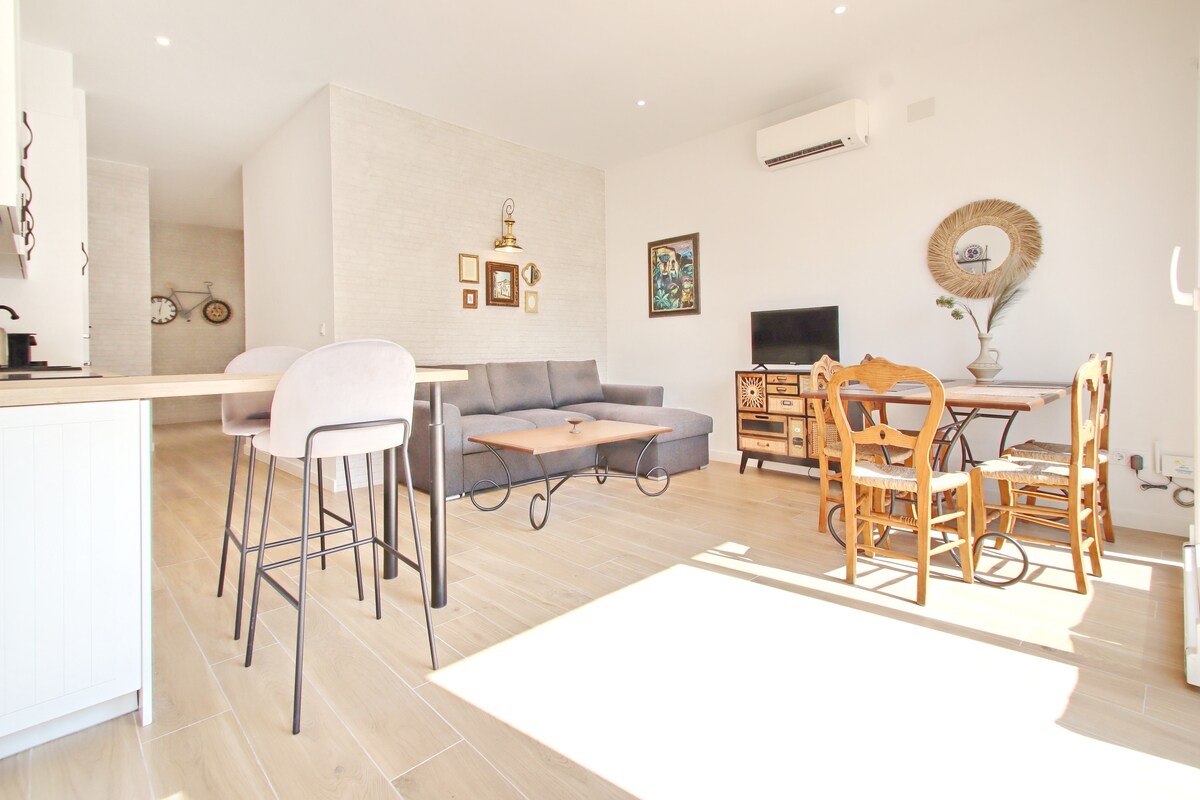 An open living area features a gray sectional sofa positioned across from a wooden coffee table. A dining set with wooden chairs is arranged beside a sideboard. Bright light fills the space from large windows, and light-colored walls enhance the airy feel.