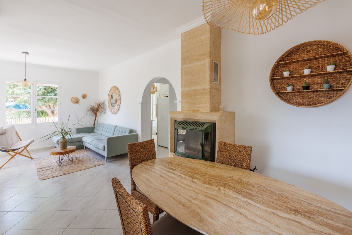 A spacious living and dining area features a light-colored stone fireplace and a wooden dining table surrounded by woven chairs. A comfortable sofa is positioned nearby, with large windows providing ample natural light and views of the garden.
