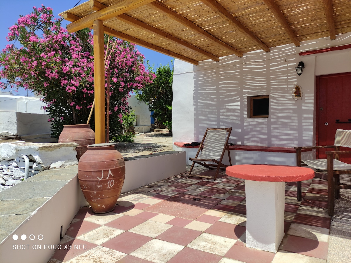 A shaded outdoor area features a rustic stone floor with a checkerboard pattern. A terracotta pot and a red-topped table are positioned beside a wooden chair. Bright bougainvillea shrubs provide a vibrant backdrop against the whitewashed wall of the structure.