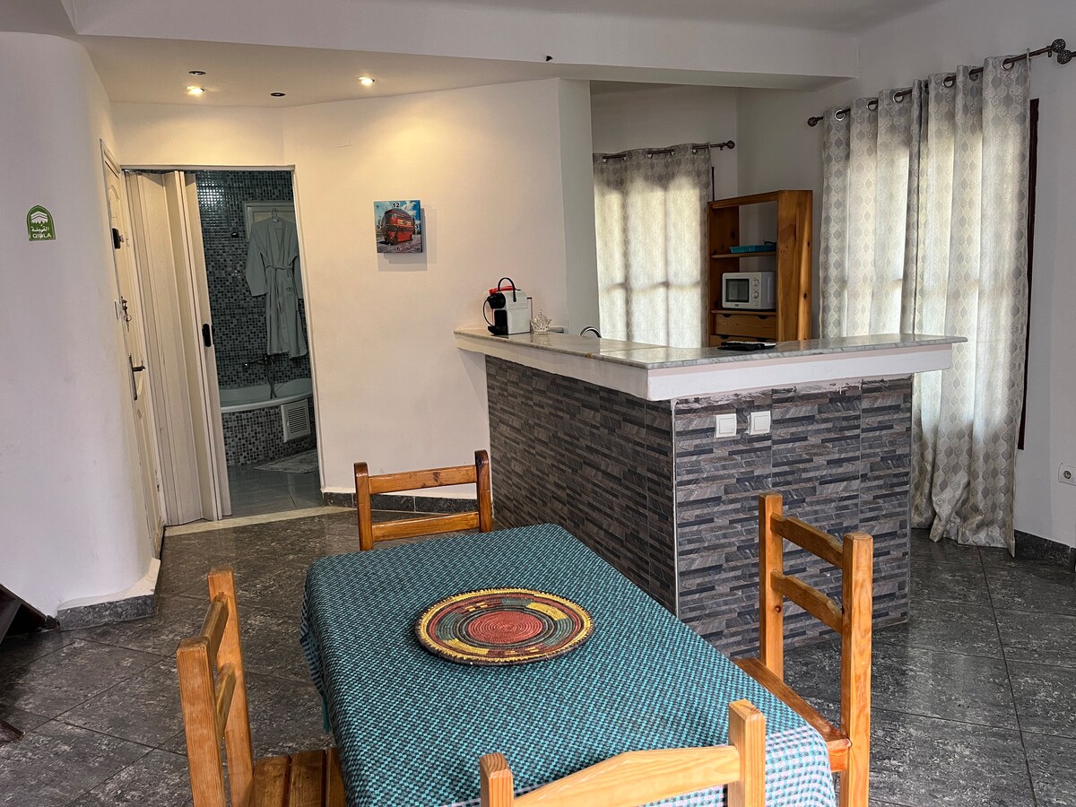 An open dining area is presented with a wooden table surrounded by four chairs, featuring a woven placemat at the center. A kitchen island with a stone facade is visible, and a microwave is placed on a wooden cabinet. Bright light enters through the large windows, partially covered by light curtains.