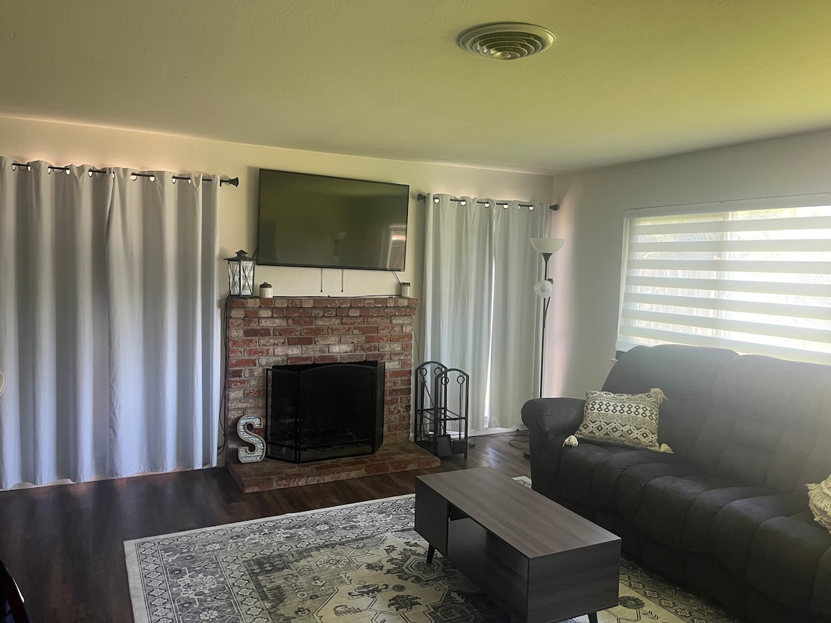 A living area features a comfortable dark sofa, centered on a low table. A flat-screen TV is mounted above a brick fireplace. Large windows with sheer drapes allow natural light to filter in, complemented by a decorative area rug placed on the hardwood floor.
