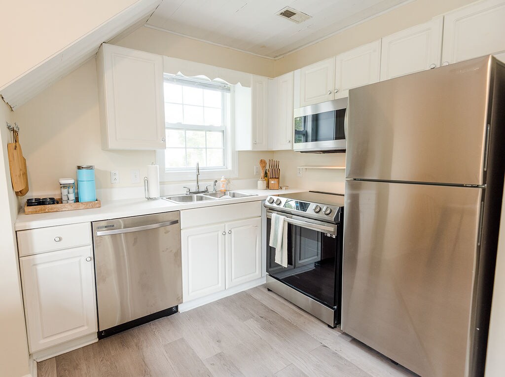 A compact kitchen features modern stainless steel appliances, including a refrigerator, oven, and dishwasher. White cabinetry provides ample storage, while a single basin sink is positioned beneath a window that allows natural light to fill the space. Neutral-colored flooring complements the design.
