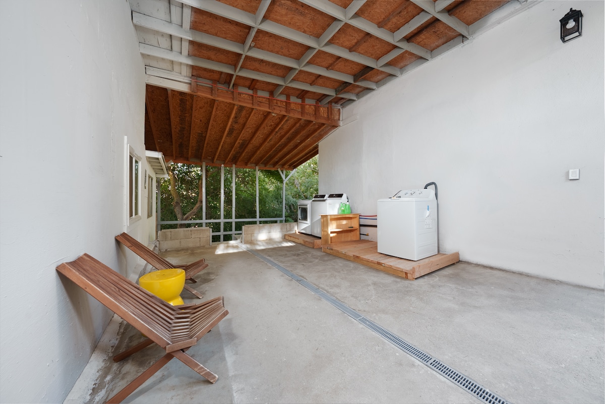 An outdoor laundry area features two washing machines on a raised wooden platform surrounded by natural greenery. Two wooden lounge chairs are positioned beside a bright yellow stool, enhancing the space's functionality. The area is covered, providing shelter while keeping an open feel.
