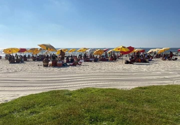A sandy beach is dotted with yellow and red umbrellas providing shade for guests. Beachgoers are seen lounging on colorful mats spread across the sand, with the blue ocean visible in the background under a clear sky.