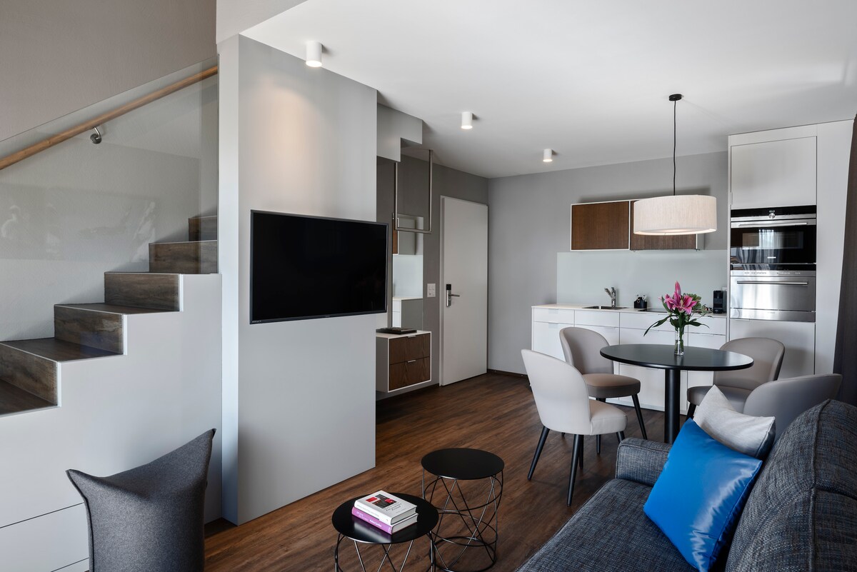 A modern living area features a comfortable sofa with a decorative pillow, a round dining table, and stylish chairs. A flat-screen TV is mounted on the wall, while a well-equipped kitchenette is visible in the background. Natural light fills the space through a nearby window.