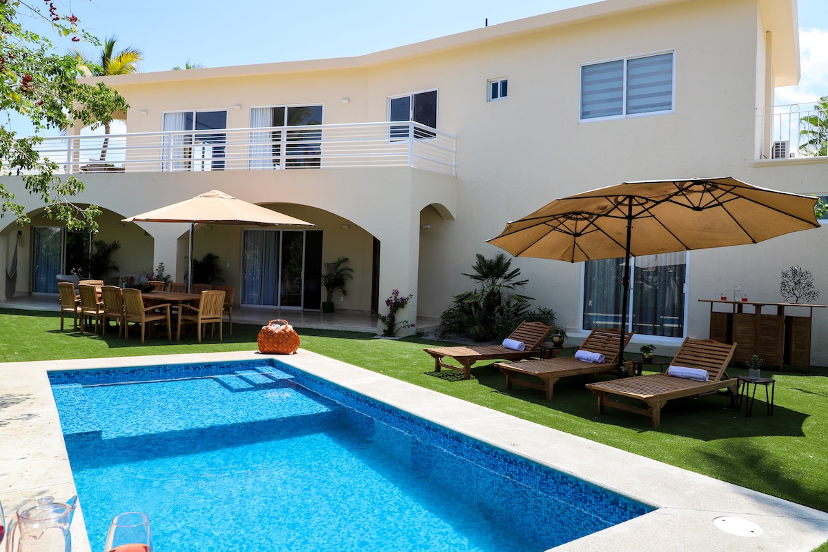 A serene outdoor area is highlighted by a private pool, surrounded by lounge chairs and shaded by large umbrellas. A dining table with chairs is positioned on the patio, while lush greenery and palm trees add to the tranquil atmosphere.