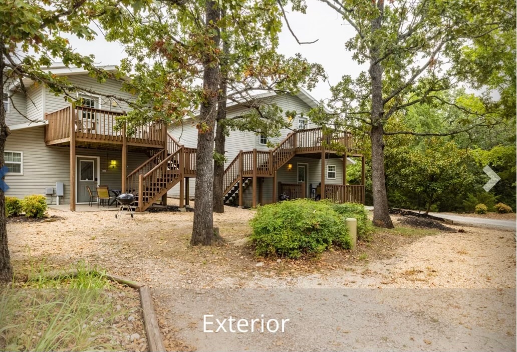 A stand-alone lodge is surrounded by mature trees, featuring two levels with wooden decks. The gravel path leads to the entrance, while outdoor seating is visible on the upper deck. The well-maintained landscape is complemented by shrubbery and natural elements.