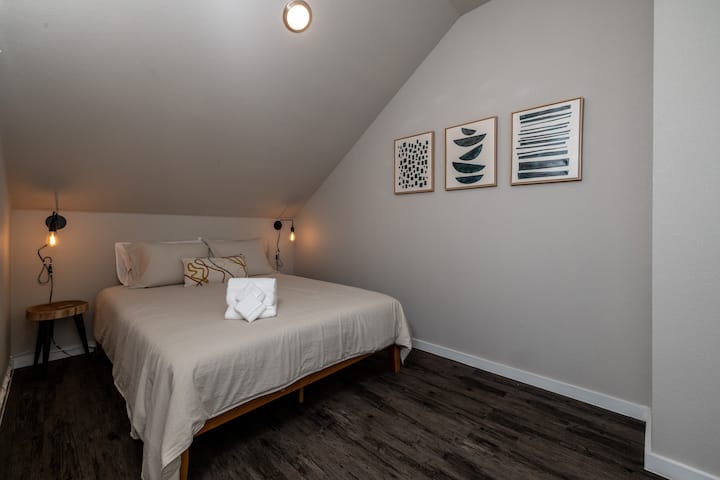 Charming fifth bedroom with a queen bed, cozy sloped ceiling, and warm hanging lights on each side—perfect for a restful, intimate vibe.  This bedroom is located upstairs.
