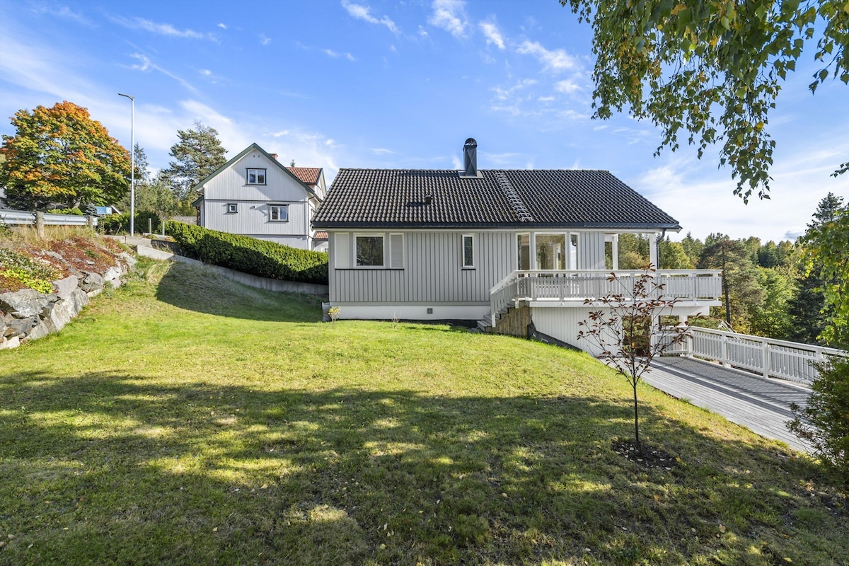 A single-story house is set in a green landscape, featuring a manicured lawn and a spacious deck. Large windows showcase views of the surroundings, while trees provide a natural backdrop. A neighboring house is visible in the distance, enhancing the tranquil atmosphere.
