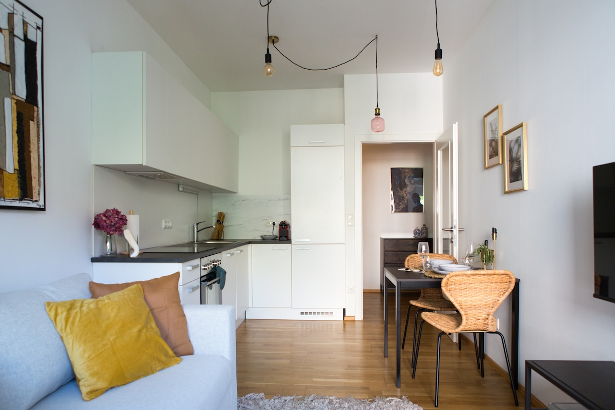 A modern living area features a light-colored sofa with a yellow cushion. A small dining table with woven chairs is positioned next to a sleek kitchen with white cabinets. Pendant lights hang from the ceiling, adding subtle illumination to the space.
