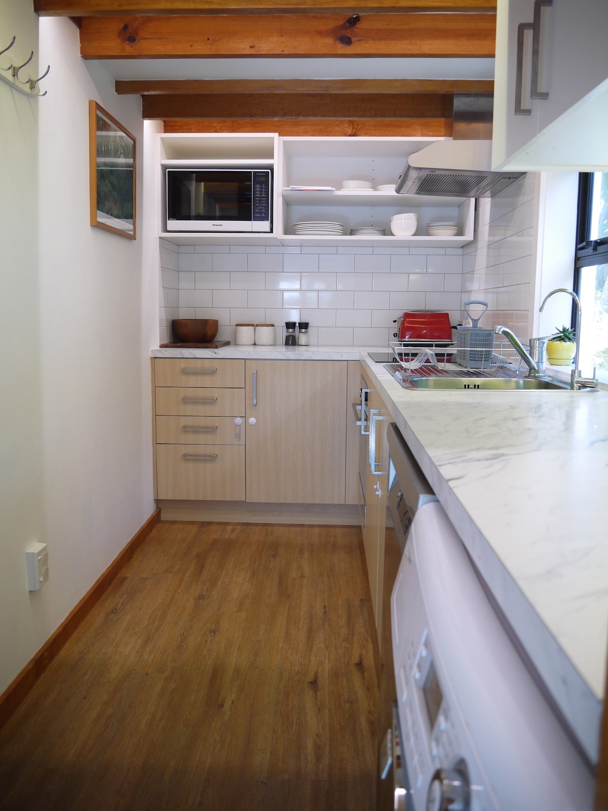 A galley-style kitchen is shown, featuring light-colored cabinetry and modern appliances. A microwave is mounted above the counter, with bowls and dishes neatly arranged above. A sink and dishwasher are present, complemented by a marble countertop and wooden flooring, enhancing the space's functionality.