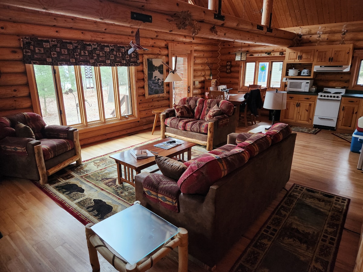 A spacious living area features two log-framed sofas adorned with patterned cushions, facing a coffee table. Large windows provide ample natural light, while a dining area can be seen in the background. The warm wooden walls and flooring create a cozy environment.
