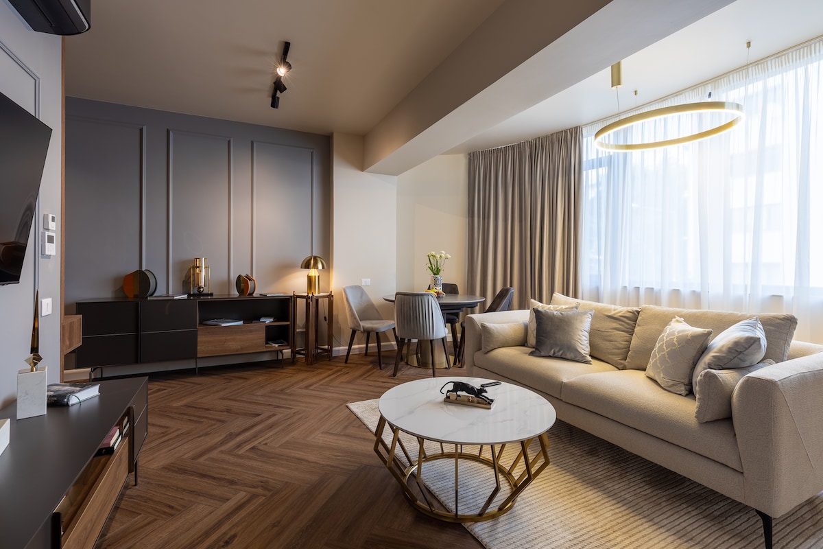 A modern living area features a soft beige sofa adorned with decorative pillows. A round marble coffee table is centered on a patterned wooden floor. Elegant lighting fixtures hang above, and large windows dressed in sheer curtains allow natural light to fill the space.