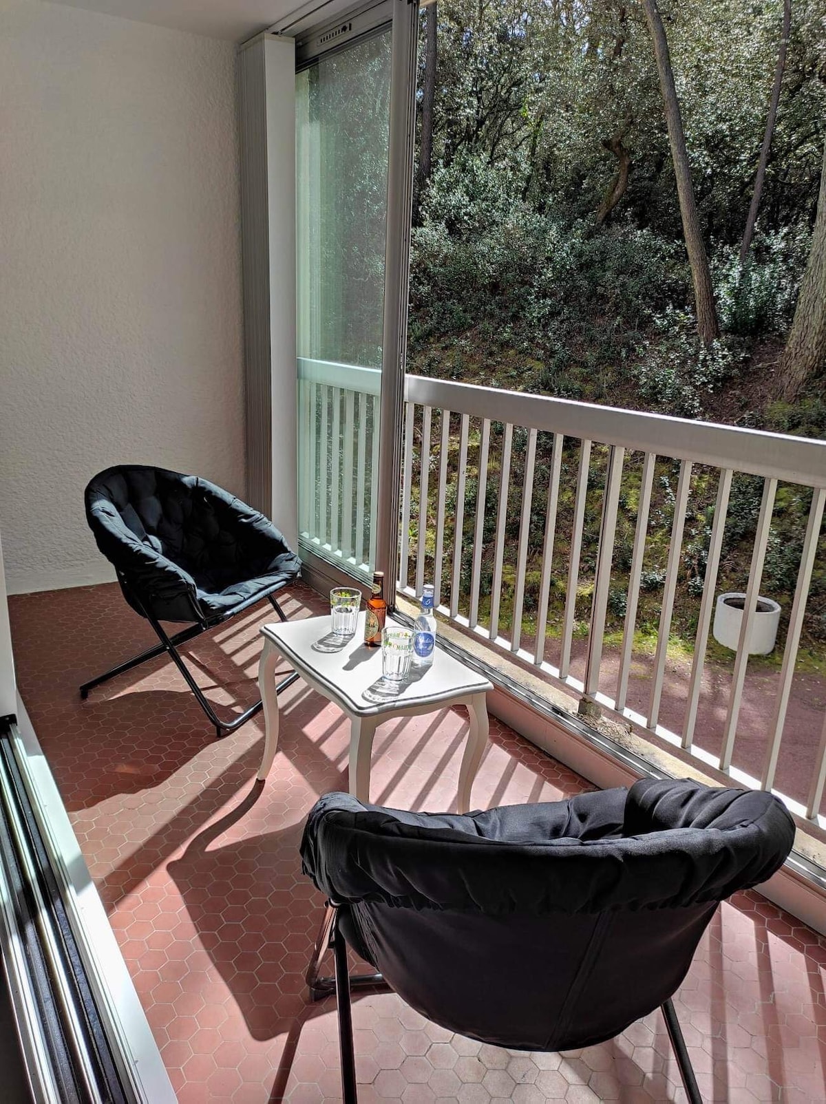 A tranquil balcony is equipped with two black chairs and a small white table. Refreshments are arranged on the table, and large glass doors provide ample natural light. The surrounding greenery enhances the peaceful ambiance, creating a serene outdoor space.
