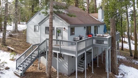 The exterior of the cabin is shown, featuring a wrap-around deck with outdoor seating and a gas grill. Tall trees surround the home, with some snow visible on the ground. Steps lead from the deck to the side of the house.