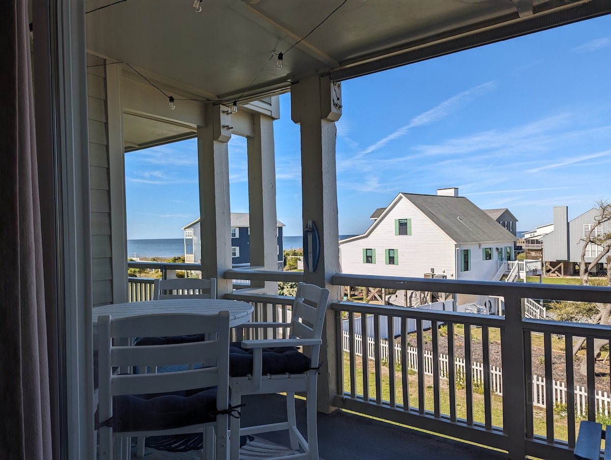 A covered balcony is visible, featuring a table with two chairs, overlooking green grass and nearby coastal homes. The ocean is seen in the distance under a clear blue sky, providing a peaceful and serene atmosphere.