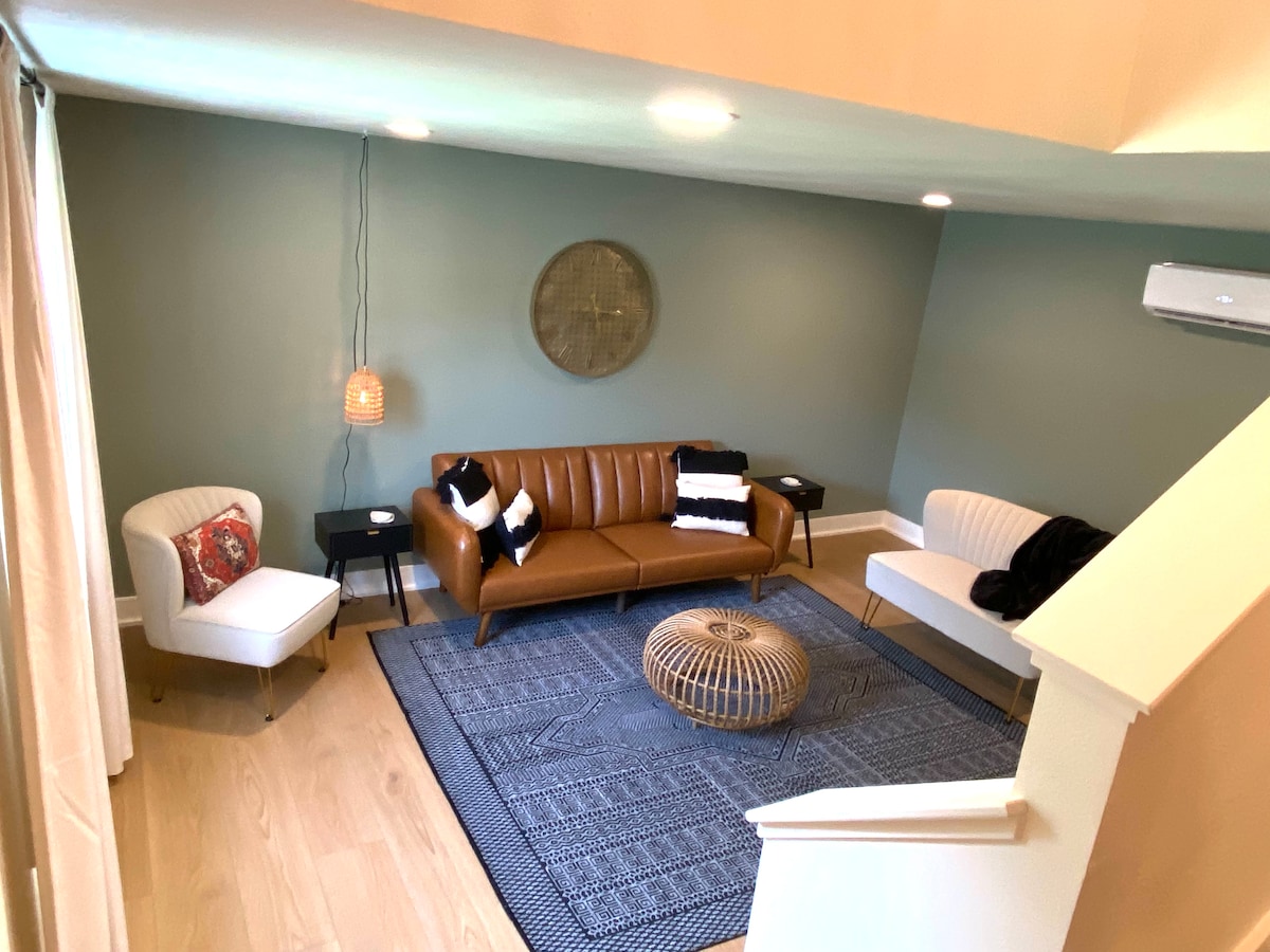 A cozy living area features a brown leather sofa paired with white accent chairs. A round, woven coffee table rests on a textured rug, and a modern light fixture hangs above. Soft lighting illuminates the mint green walls, creating a relaxed ambiance.