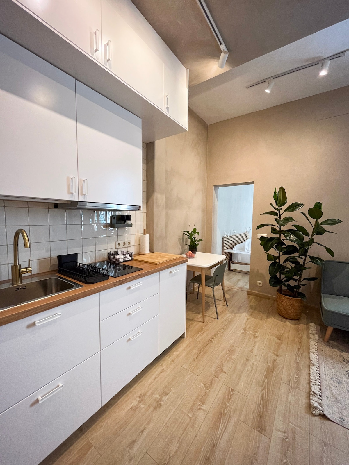 A modern kitchen area features sleek white cabinets and a wooden countertop with a gold faucet. A dining table with chairs is positioned nearby, accompanied by a tall indoor plant. Soft lighting enhances the space, leading to an inviting view of the adjacent room.