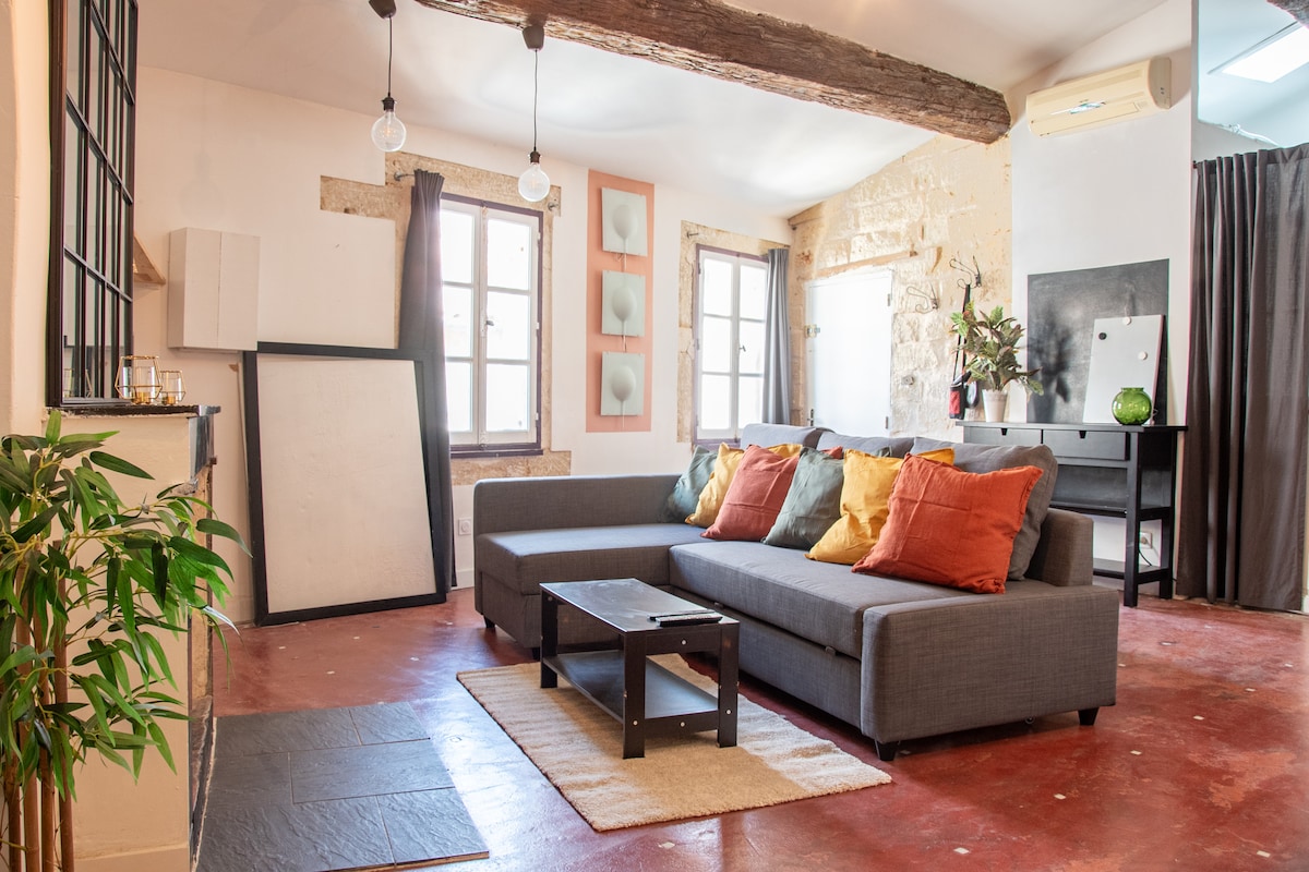 A spacious living area features a dark grey sectional sofa adorned with colorful throw pillows, complemented by a wooden coffee table. Large windows provide natural light, while exposed wooden beams add character to the ceiling. A rug is placed on the red-tinted floor.