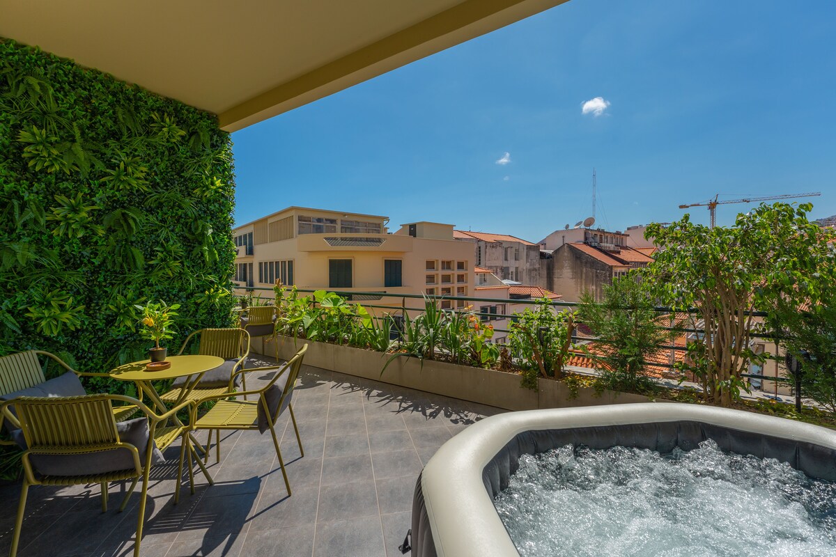 A balcony is shown with a hot tub surrounded by greenery, offering a view of the city skyline. A circular table and four chairs are positioned nearby, enhancing the outdoor space with natural elements and urban scenery.