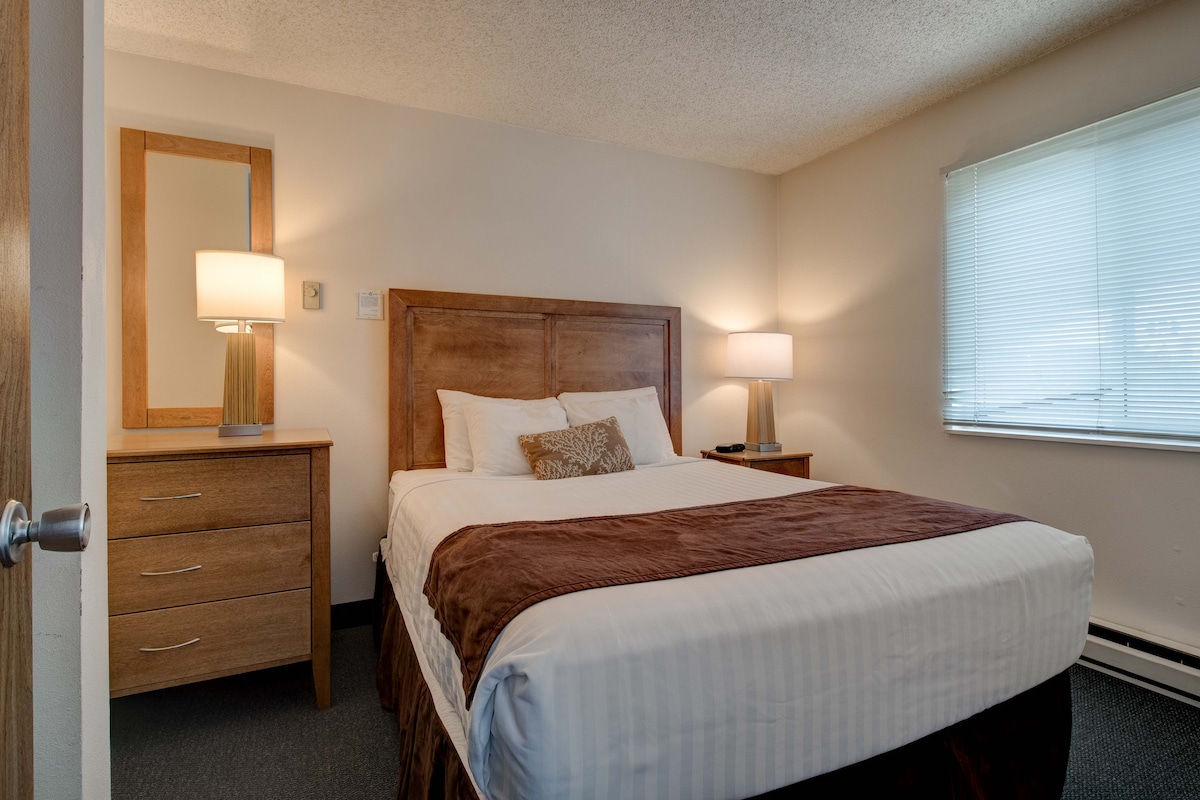 A comfortable sleeping area is highlighted by a wooden bed frame with a cozy blanket draped across. Nightstands with lamps provide soft lighting, and a dresser adds storage space. Natural light filters through the window, which has partially closed blinds.