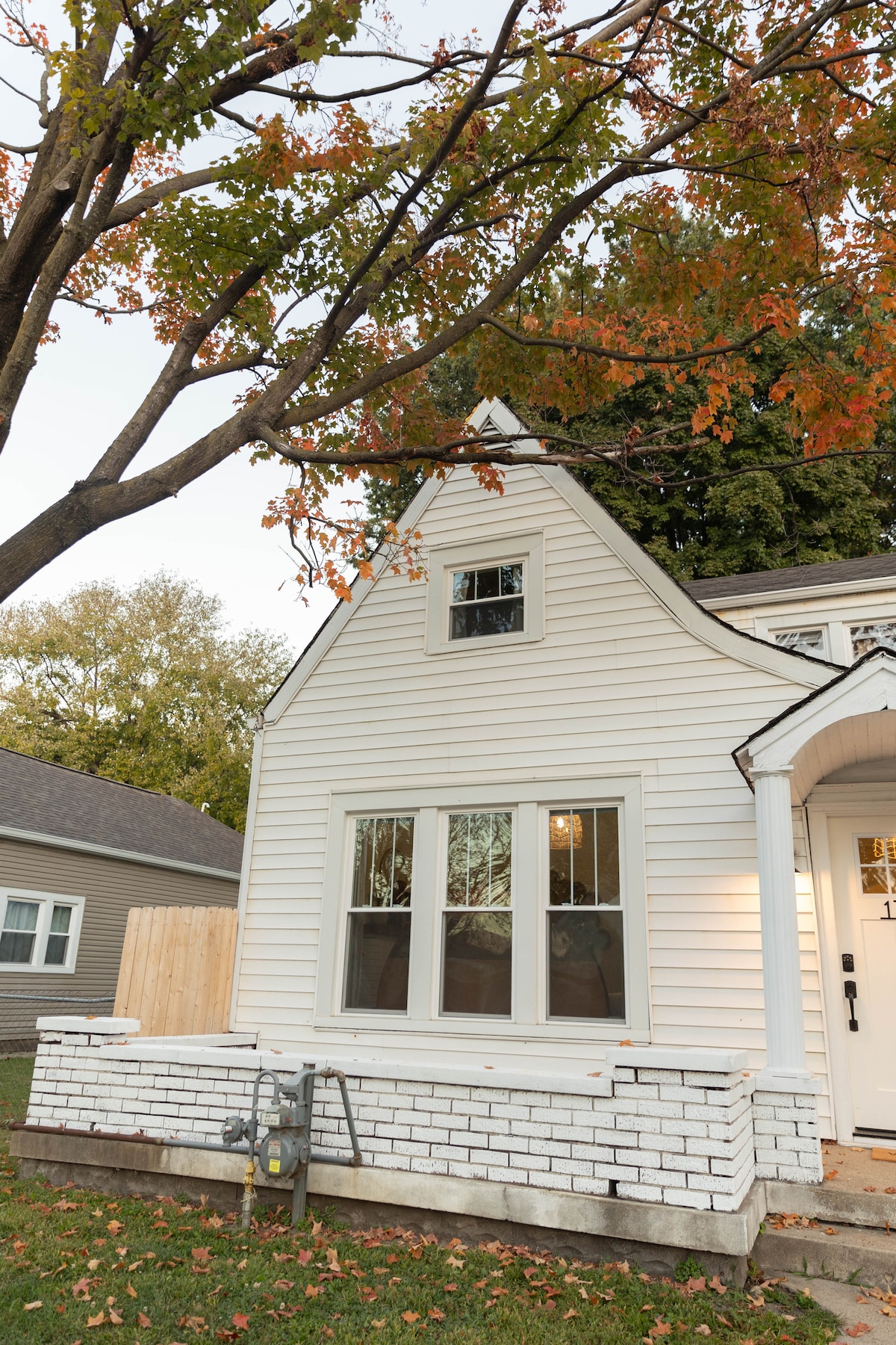 The charming exterior of the cottage showcases a gabled roof and white clapboard siding. Large windows offer a view into the living space. The front porch is bordered by a clean white brick foundation, and a tree with autumn leaves provides a splash of color.