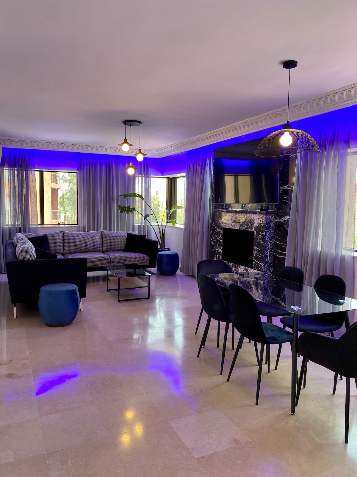 An open living and dining area is presented, featuring a sleek black sofa and a marbled fireplace. Large windows allow natural light, complemented by soft blue lighting. A glass dining table with black chairs is positioned centrally, enhancing the spacious layout.