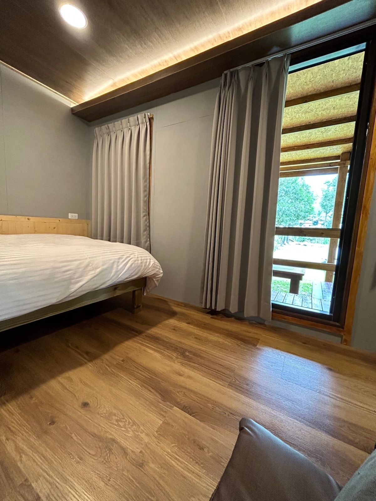 A cozy bedroom is highlighted by a spacious bed with crisp white linens. Natural light enters through a large sliding door, which leads to an outdoor space. Soft gray curtains frame the doorway, while warm wood flooring accentuates the inviting atmosphere.