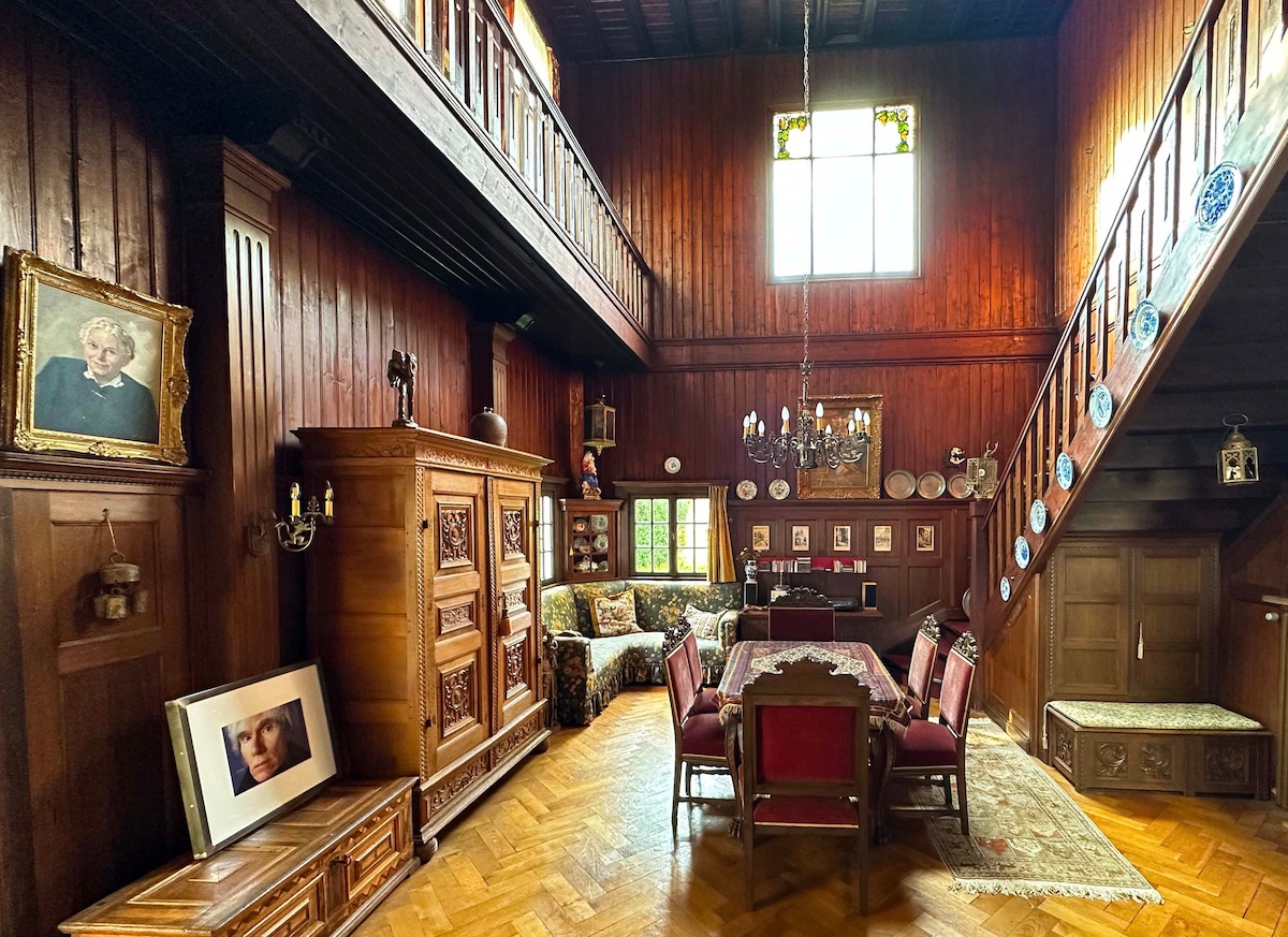 A wooden interior space features high ceilings with exposed beams and large windows that invite natural light. A dining area with dark wooden furniture is complemented by a traditional chandelier. Ornate decorations and a mix of antique and modern elements create an eclectic ambiance.
