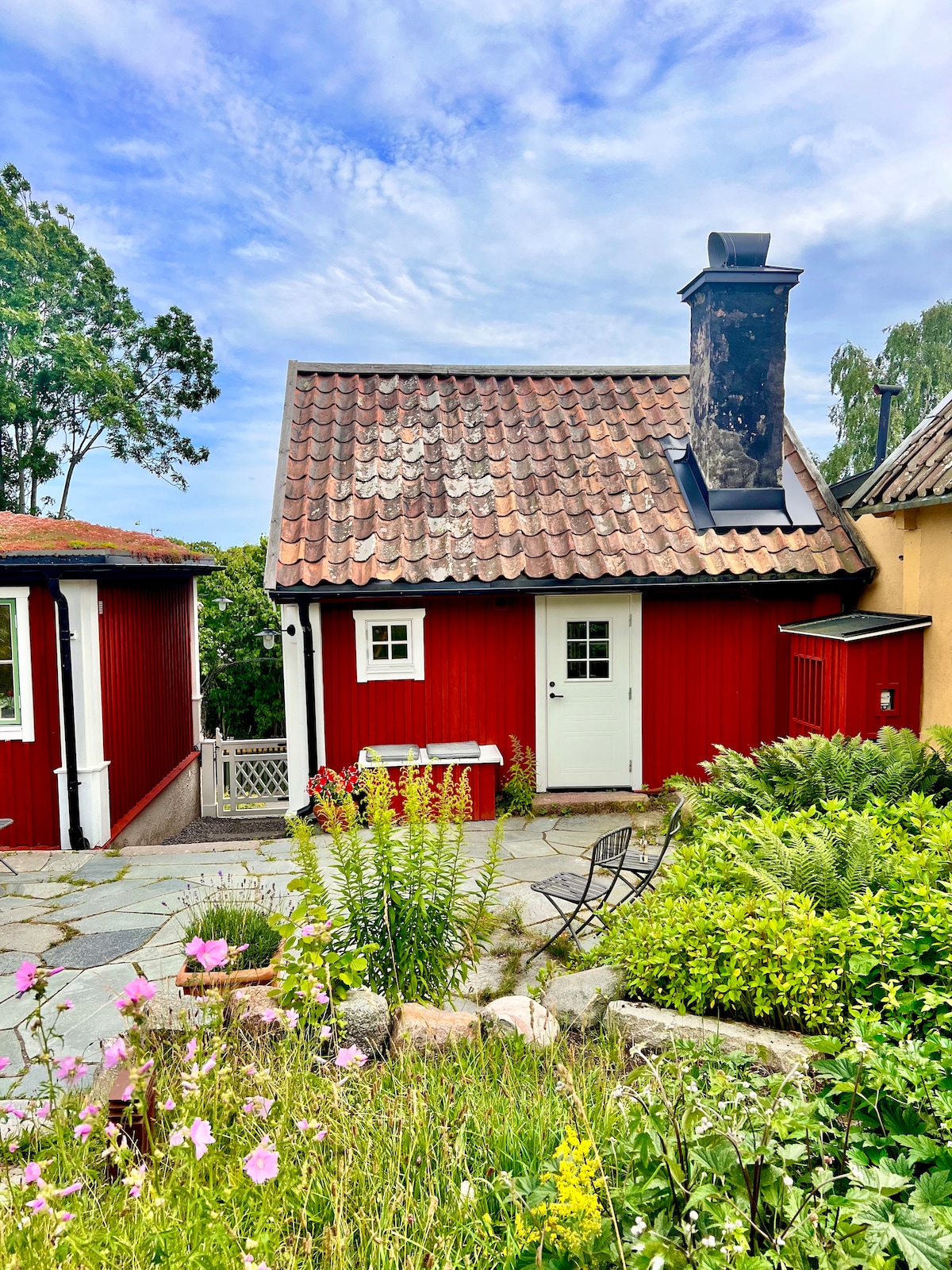 A charming red cottage with a tiled roof is framed by lush greenery and blooming flowers. A small stone patio leads to the entrance, featuring a white door and a window. The serene atmosphere is enhanced by the surrounding natural landscape.
