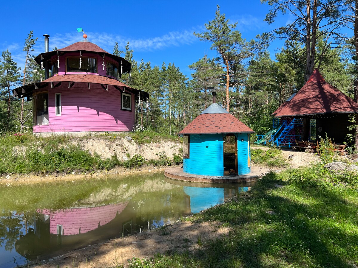 A colorful round structure and a unique blue sauna are situated by a tranquil pond, surrounded by lush pine trees. The structures feature distinct roofs and vibrant exteriors, with the sauna designed for relaxation after pond activities, offering a serene escape in nature.