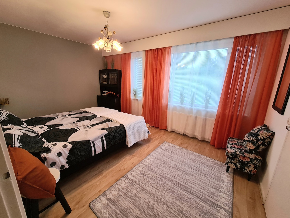 A well-arranged bedroom features a spacious bed dressed in patterned linens. Large windows with sheer curtains allow natural light to filter in. A tasteful area rug covers the wooden floor, while a small chair adds a touch of character to the room.