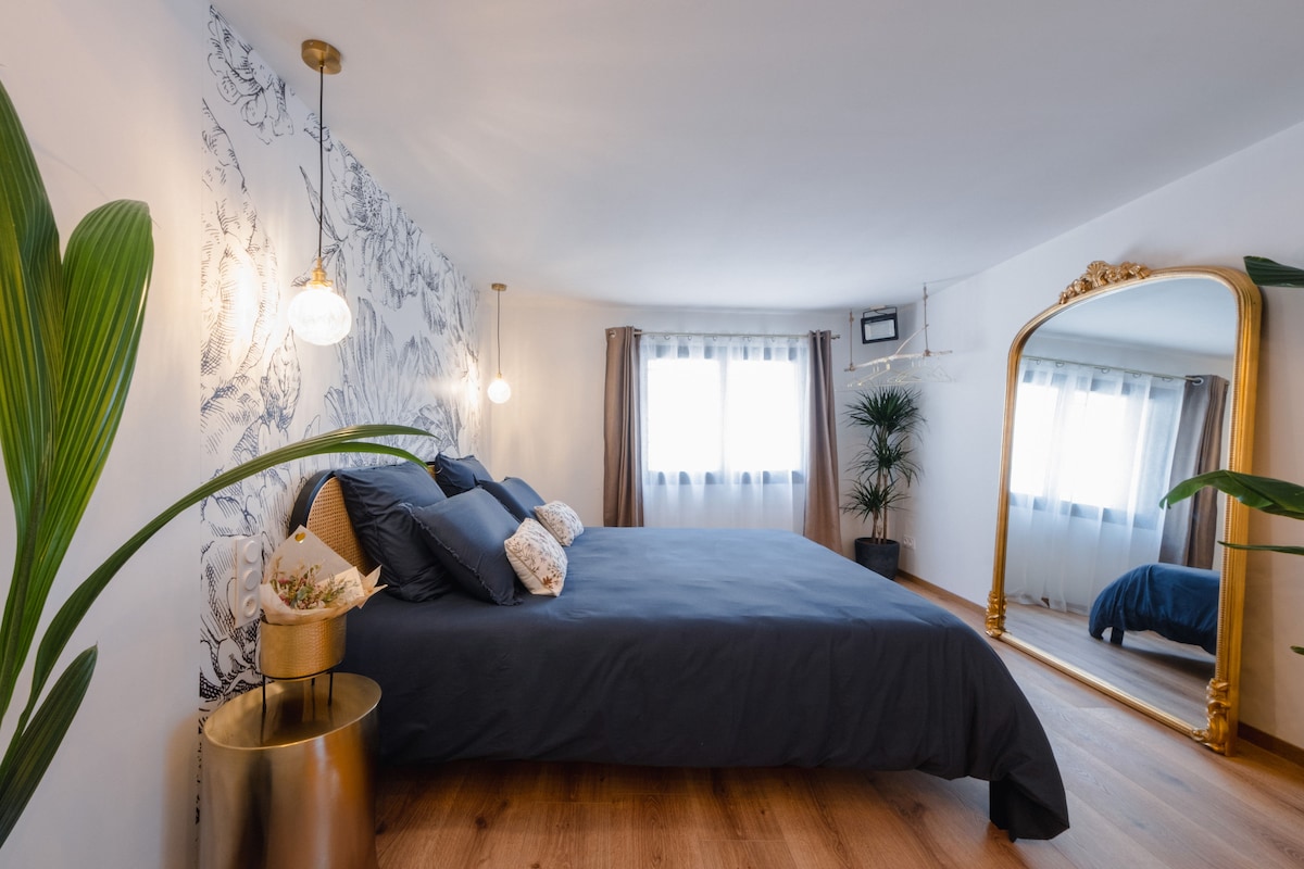 A spacious bedroom features a large dark bed with decorative pillows, complemented by an elegant golden mirror. Natural light filters through sheer curtains, illuminating the room. A plant adds a touch of greenery, enhancing the serene atmosphere.