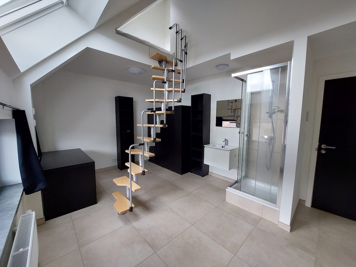 A modern room is showcased with light-colored tile flooring and neutral walls. A spiral staircase leads to a loft area. In one corner, a glass-enclosed shower is visible, accompanied by a white vanity. Large storage units stand along the walls, creating a spacious environment.
