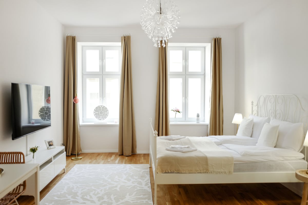 A spacious bedroom is highlighted by a large window allowing natural light to fill the room. A stylish chandelier hangs above the bed, which is neatly dressed in white linens. A television is mounted on the wall opposite a light-colored sideboard.