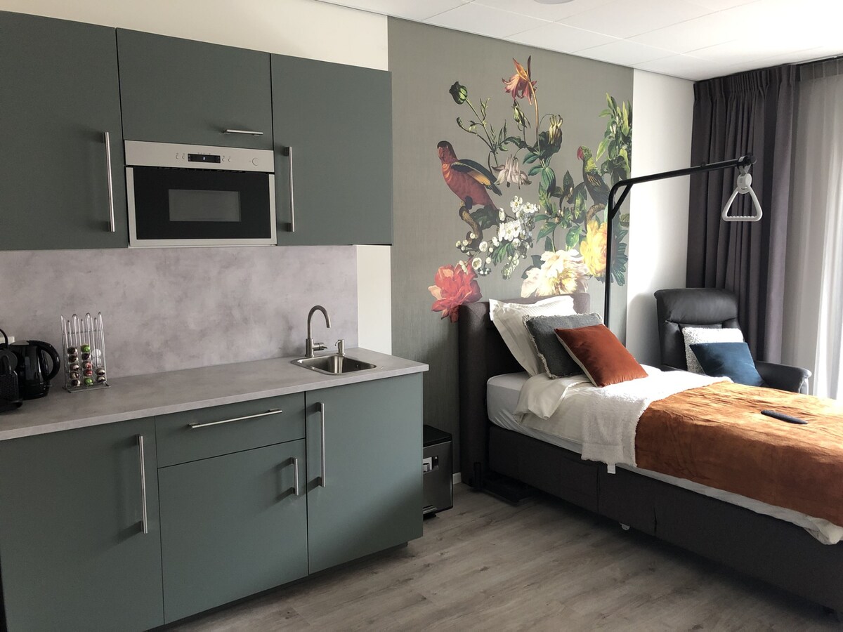 The room features a kitchenette with a microwave and sink, complemented by a floral wall mural. A comfortable bed with decorative cushions sits adjacent to a chair. Light filters through the floor-to-ceiling window, accented by soft drapes.